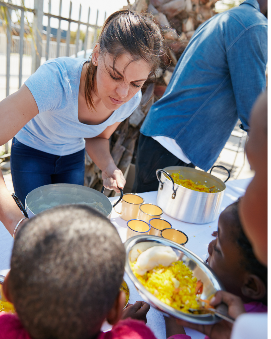 A woman serving food at an outdoor gathering with children. There are pots of rice and a plate of rice with a scoop in a colorful setting.
