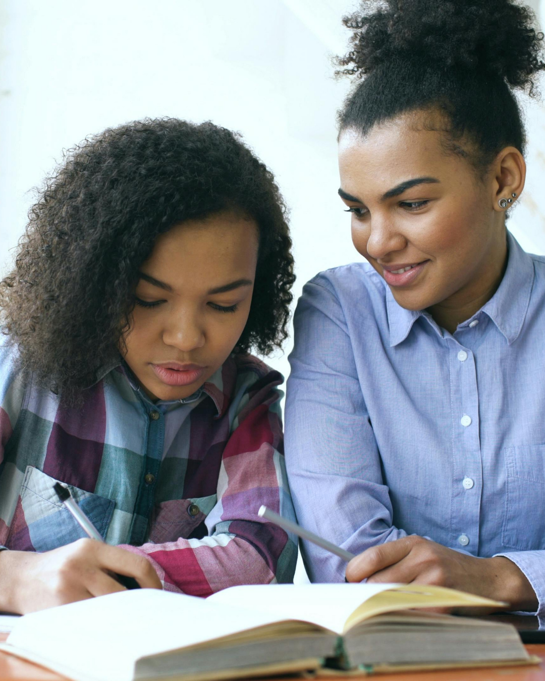Two women studying together, one writing in a notebook and the other observing, in a classroom setting.