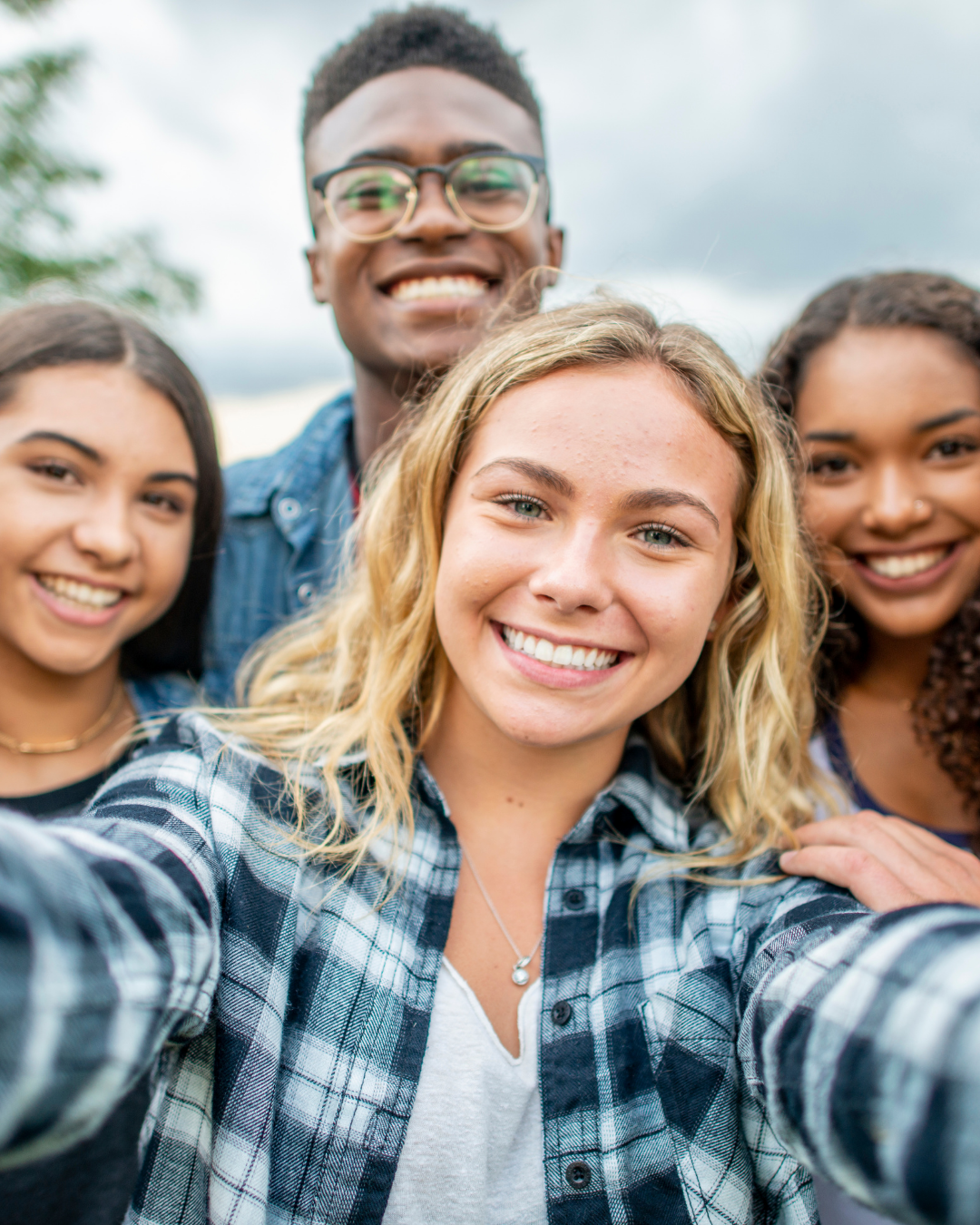 Group of four friends taking a selfie outdoors on a cloudy day, smiling and close together.