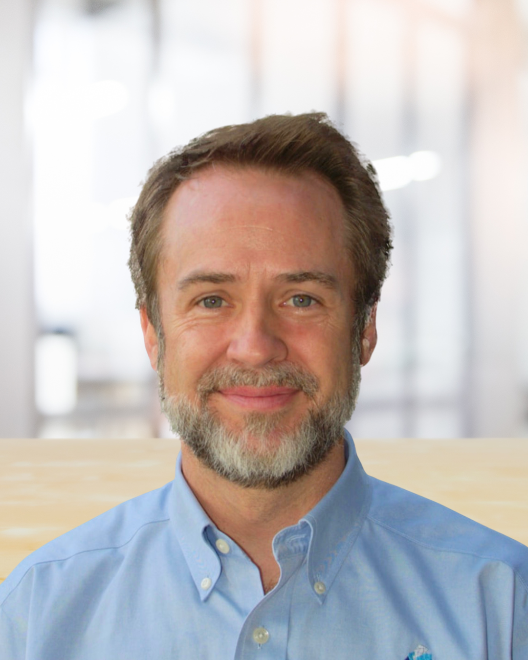 Portrait of a middle-aged man with brown hair and a beard, wearing a light blue button-down shirt, smiling in a bright, modern office setting.