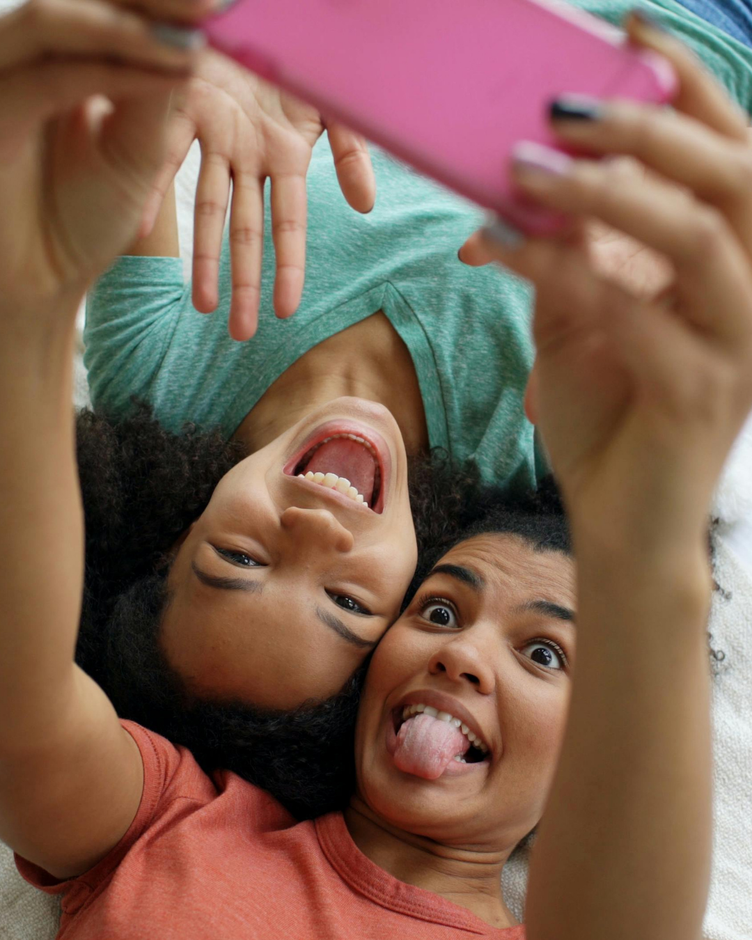 Two women lying down, taking a selfie together, making funny faces with tongues out and smiling.