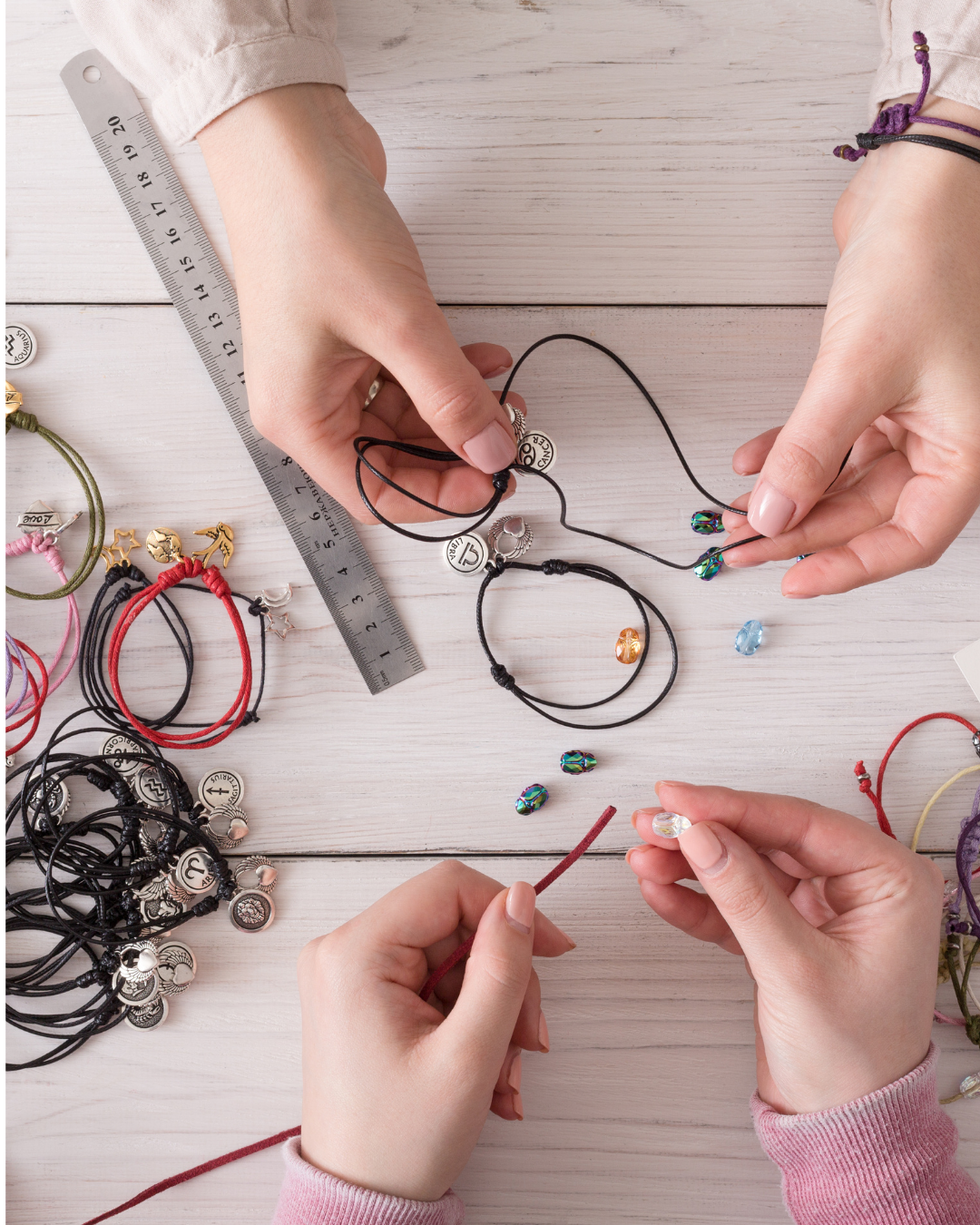 Multiple hands working on creating jewelry with colorful beads, charms, and cord on a white wooden surface, including a metal ruler for measuring.