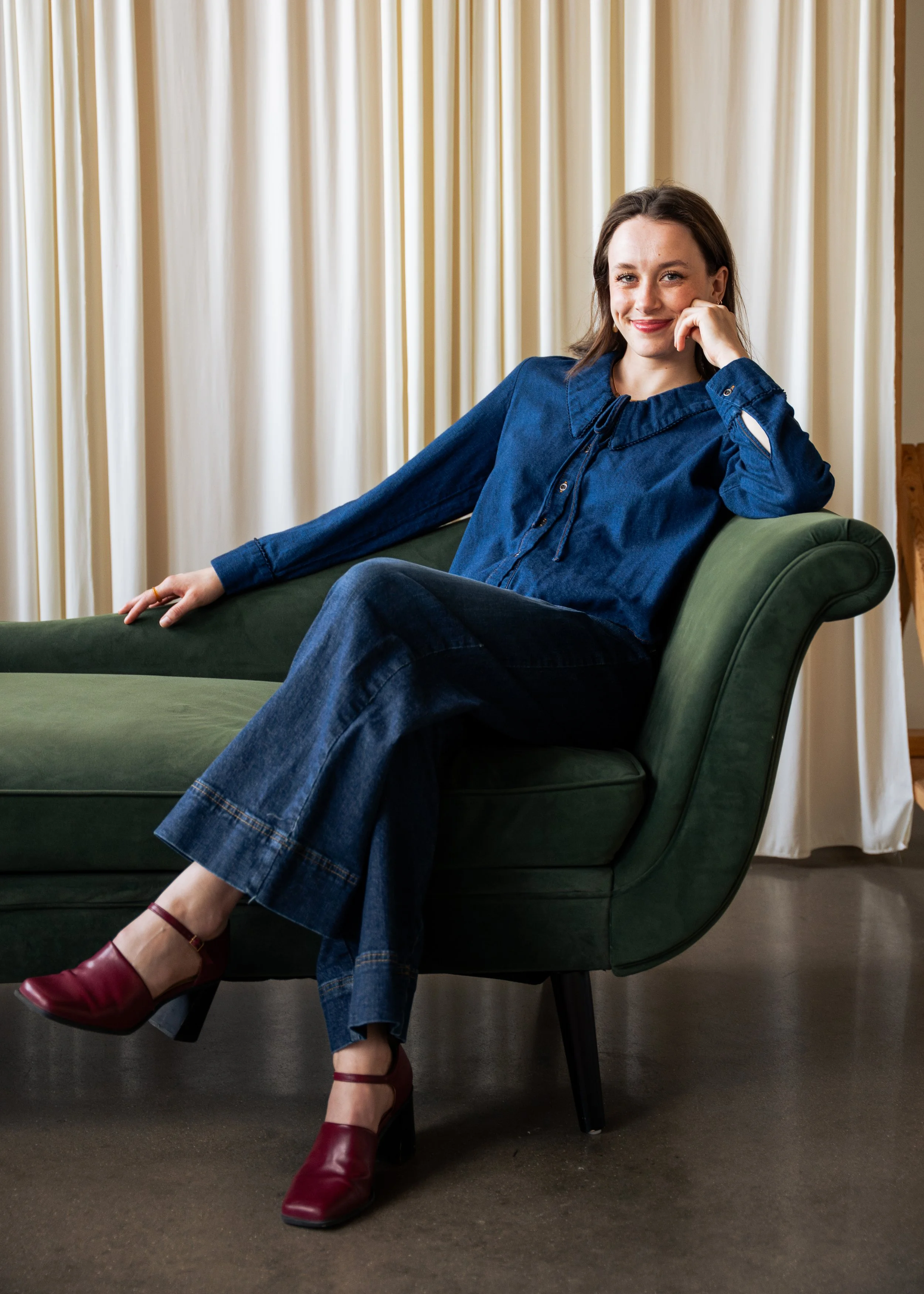A woman sitting on a green couch with a cream curtain background, wearing a denim outfit and red high heel shoes, smiling and resting her head on her hand.