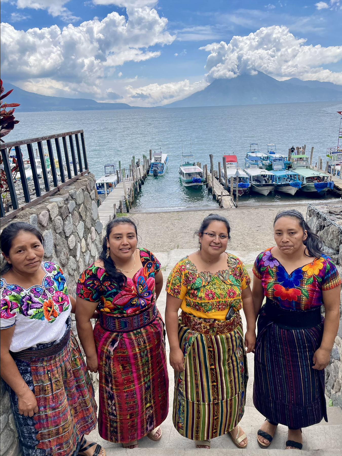 Four women standing in front of Lake Atitlán with boats docked at the pier, lush mountains and Volcano Atitlán in the background, all wearing colorful traditional Mayan clothing.