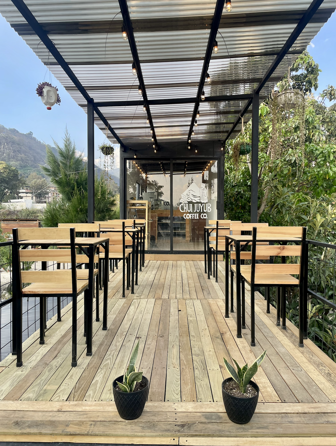 Outdoor terrace of a coffee shop with wooden tables and chairs, potted plants, string lights, and a glass door with the cafe's logo, surrounded by greenery and mountains in the background.