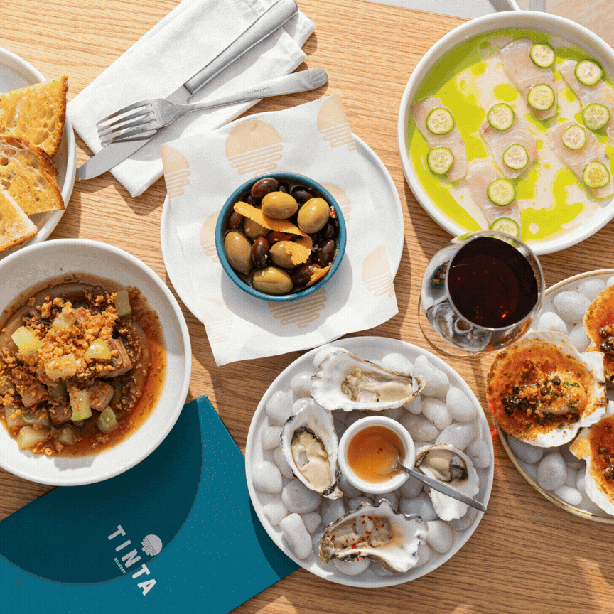 A variety of dishes on a wooden table, including oysters with lemon wedges, a bowl of marinated olives, a plate of sliced bread, a bowl of soup with vegetables, a glass of red wine, and a dish of baked oysters.