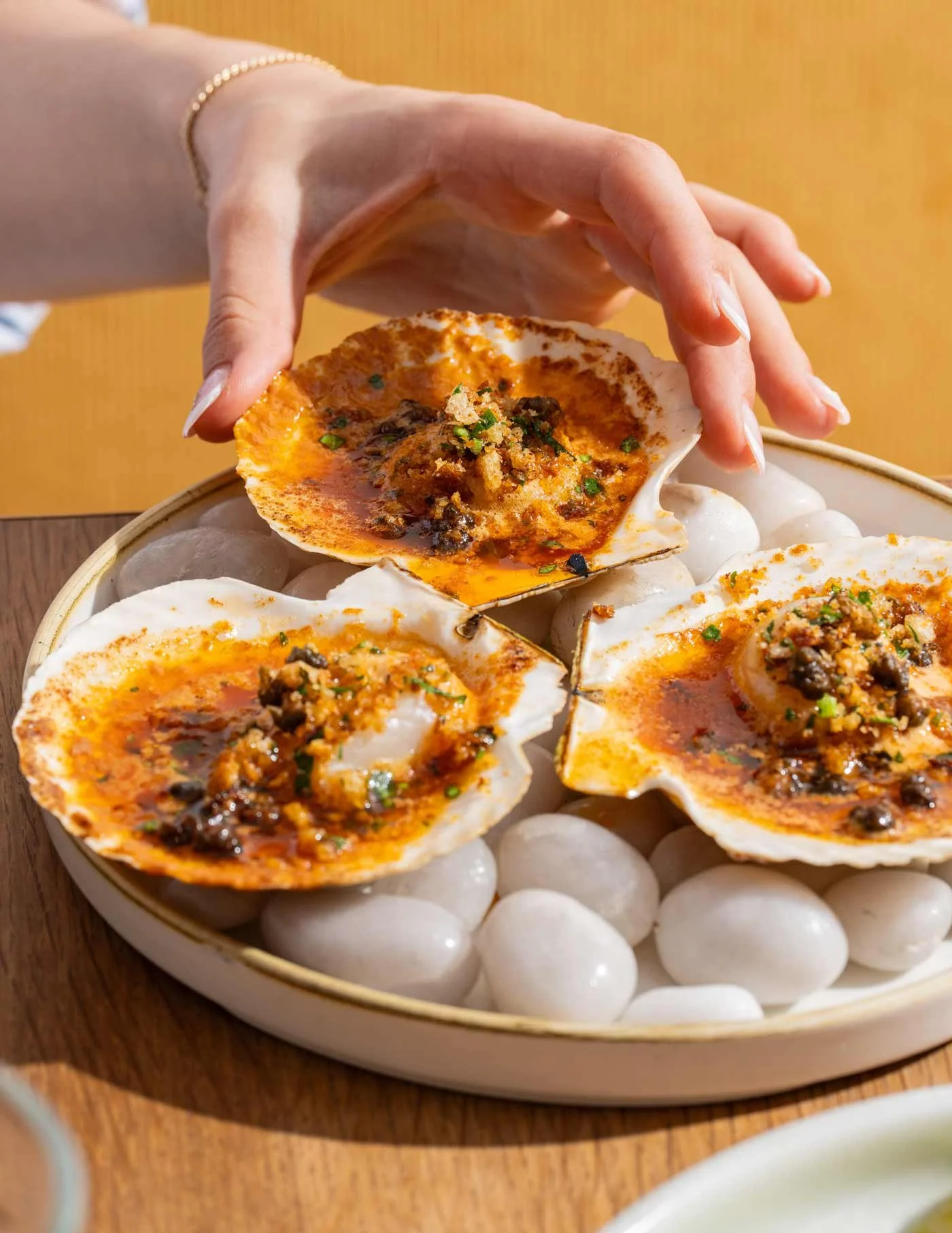 A hand wearing a gold bracelet holding a scallop shell with spicy topping over a plate of similar scallops resting on white stones.