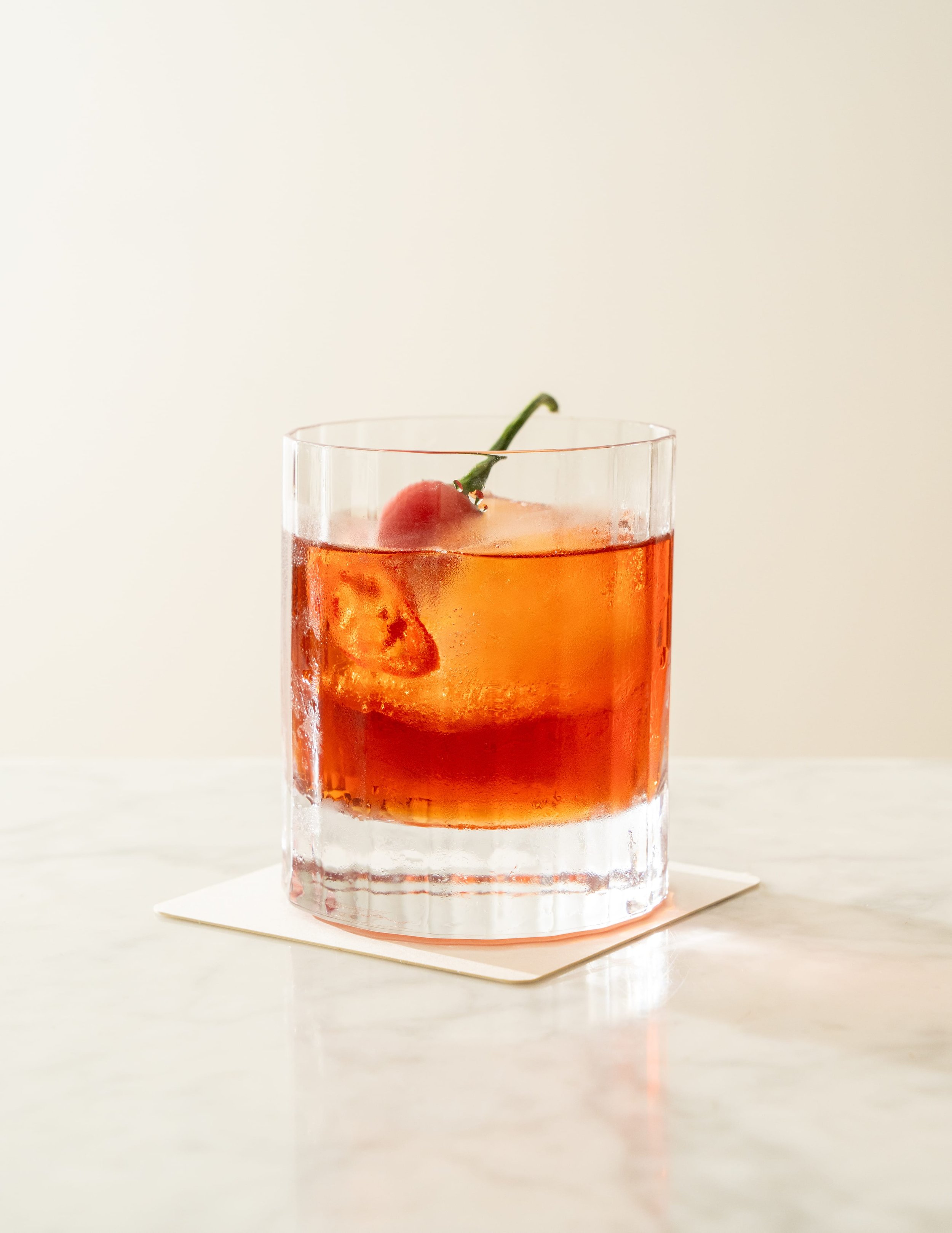 A clear glass of orange-colored beverage with a cherry and a slice of fruit inside, placed on a white surface against a plain background.