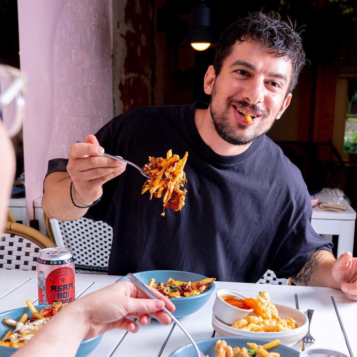 A man with curly dark hair and a beard smiling while holding a fork with pasta and sauce, sitting at a table with other bowls of pasta, a hand reaching with a fork, and a can of beer in an indoor dining area with dim lighting.