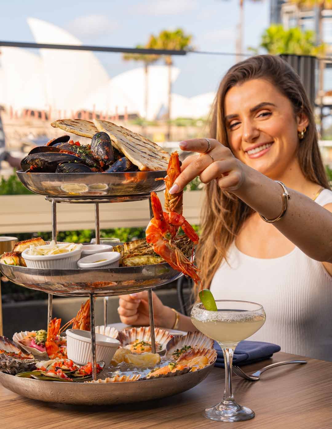 A woman smiling and holding a cooked shrimp at an outdoor restaurant with a three-tiered seafood platter and a cocktail on the table.