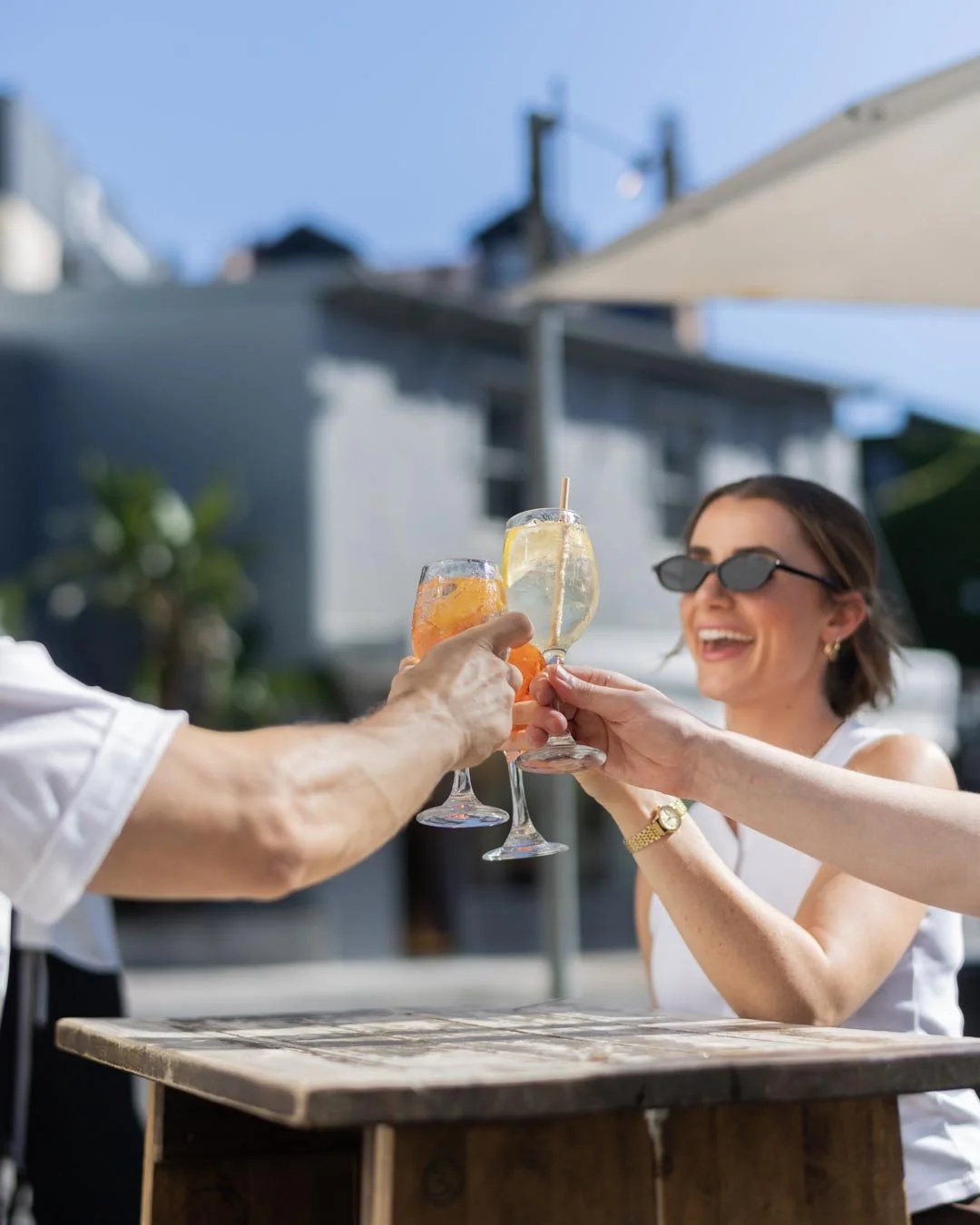 People clinking glasses of cocktails outdoors during daytime, smiling, with a woman wearing sunglasses.