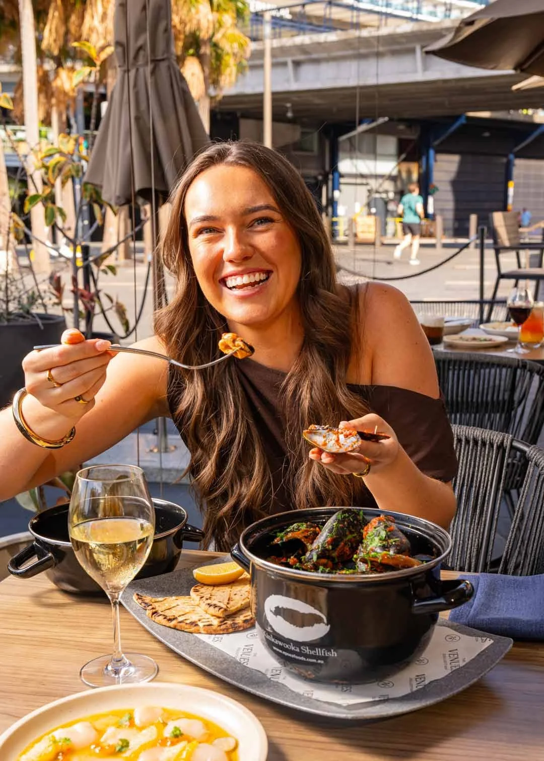 A smiling woman with long brown hair enjoying a meal at an outdoor restaurant, with a black pot of seafood, a glass of white wine, and a plate of grilled bread on the table. The background shows some other people and a street scene.