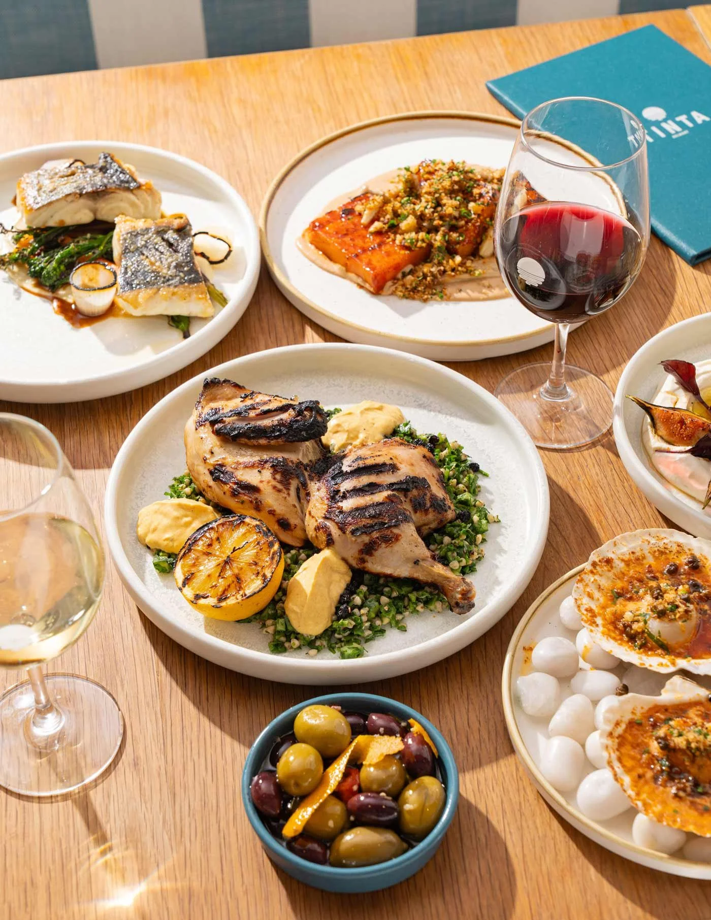 A table with various dishes including roasted chicken with lemon and herbs, grilled fish with a sauce, pasta with tomato sauce, a bowl of mixed olives, a plate of tapas with clams and sauce, and a glass of red wine.