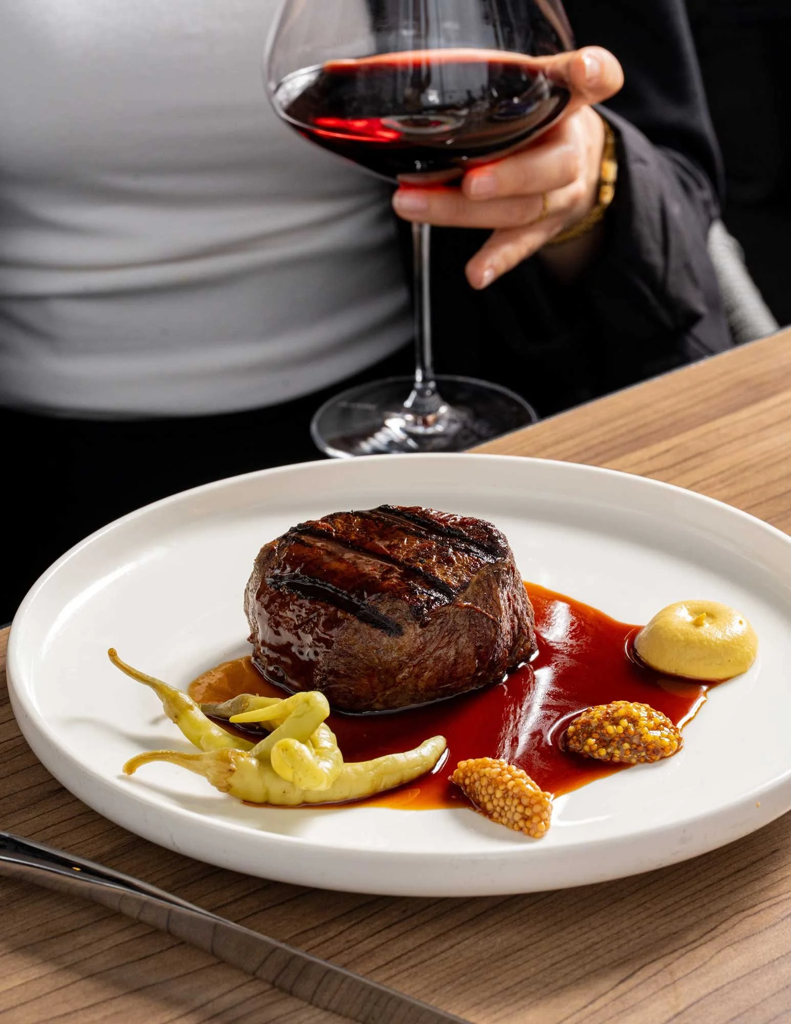 A plated serving of steak with sauce, garnished with vegetables and mustard, with a glass of red wine in the background.