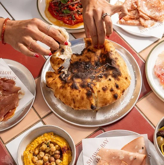 Person tearing a piece of burnt pizza with hands, surrounded by plates of prosciutto, chickpea hummus, sliced meats, and tomato salad on a tiled table.