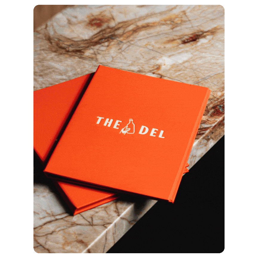 Orange menu or notebook with the text "THE DEL" and a beer bottle illustration, placed on a marble surface.