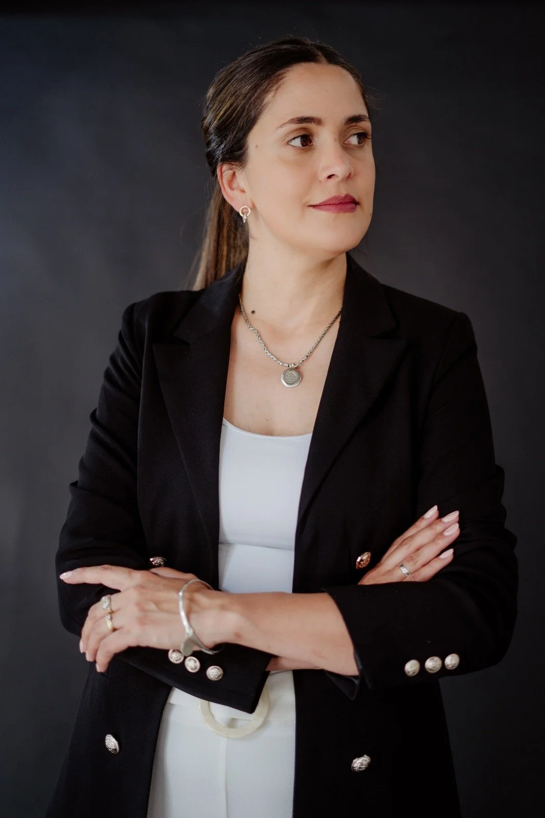 Mujer adulta con cabello largo y oscuro, vestida con saco negro y blusa blanca, lleva accesorios de joyería plateados, posando con brazos cruzados contra fondo oscuro.