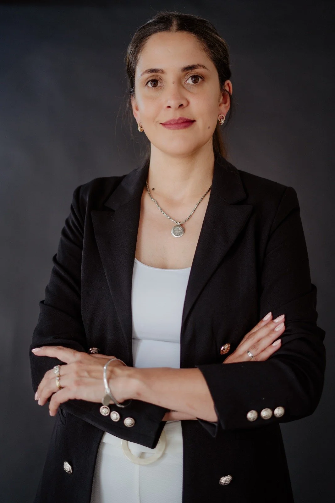 Retrato de una mujer con cabello oscuro y ojos marrones, vestida con saco negro, camisa blanca y accesorios plateados, posando con los brazos cruzados contra un fondo gris oscuro.