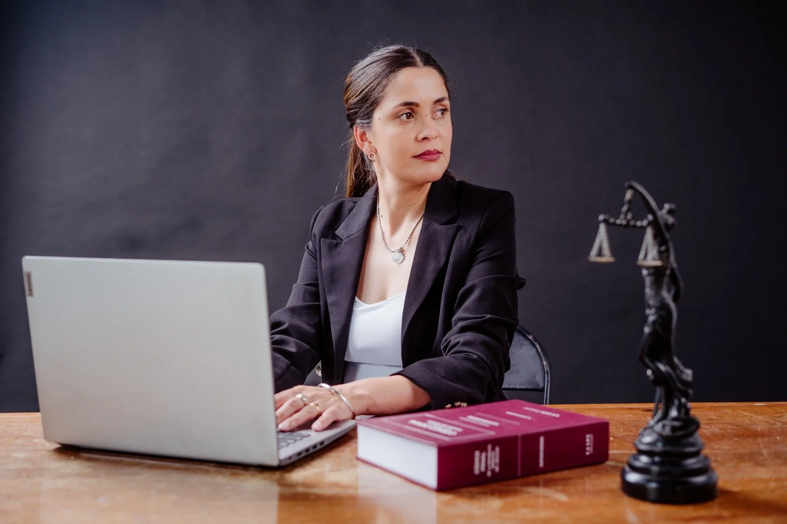 Mujer sentada en una mesa con una laptop, livros y una estatua de la justicia, vista de perfil, fondo negro, vestida formalmente con un saco negro y blusa blanca.