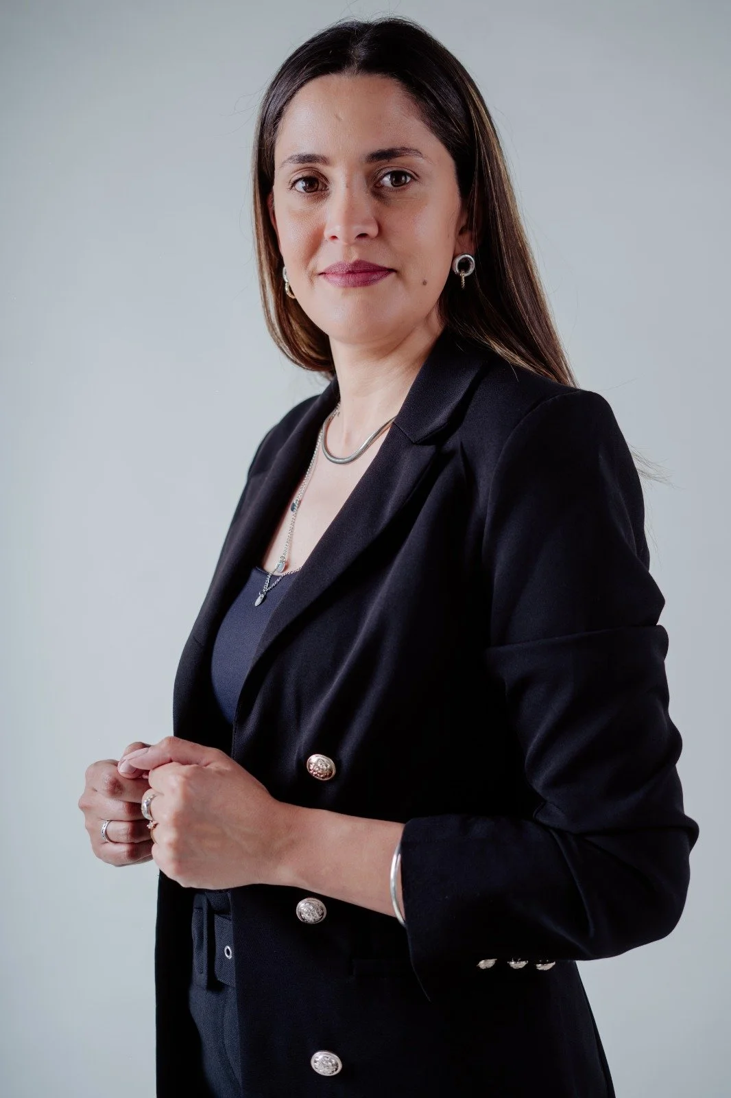 Retrato de una mujer vestida con traje negro, con cabello largo y oscuro, usando joyas y con fondo gris claro.