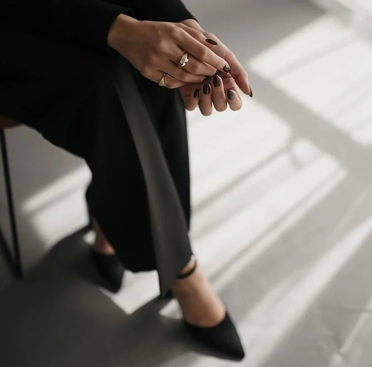 Manos de mujer con uñas pintadas de negro, usando anillos, en posición de reposo. Ella lleva un vestido negro y zapatos negros, sentada en una silla. La luz natural crea sombras en el suelo.