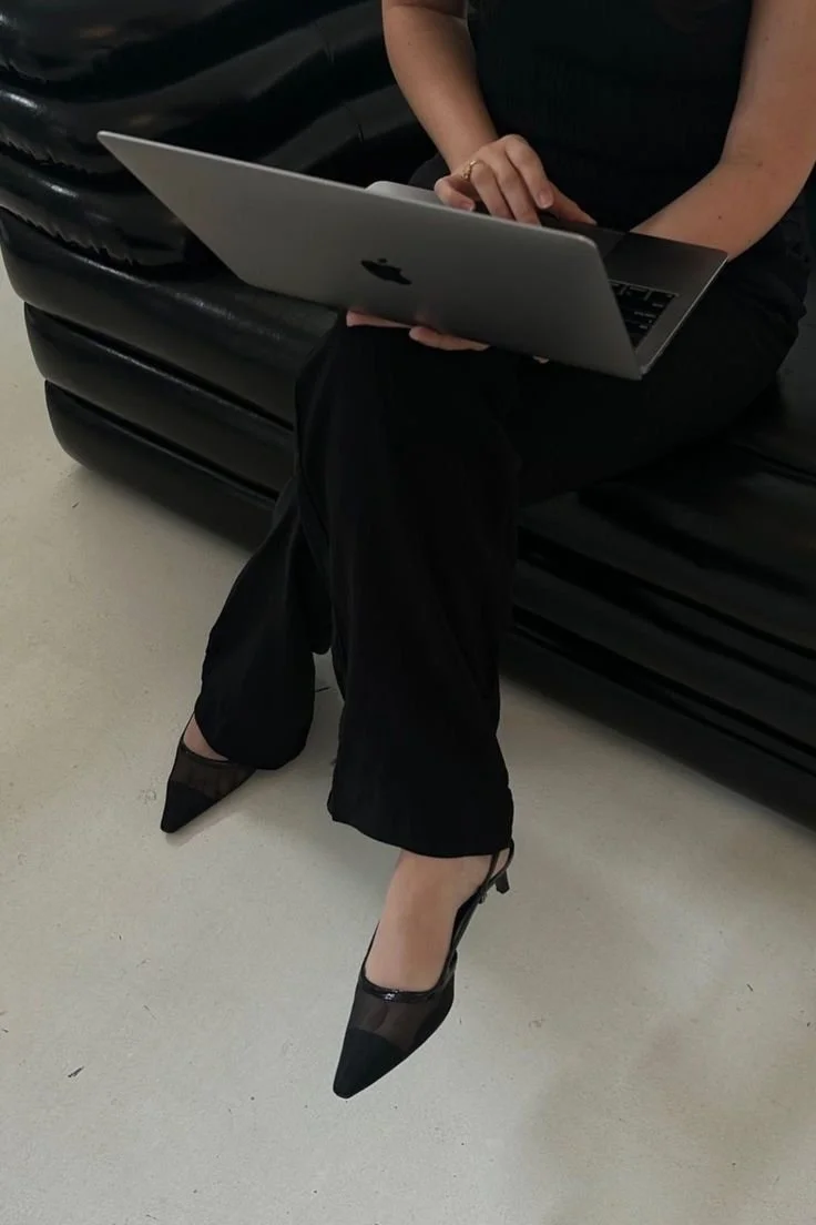 Persona sentada en un sillón negro, usando zapatos de tacón negros con detalles de malla, sosteniendo una computadora portátil en su regazo.