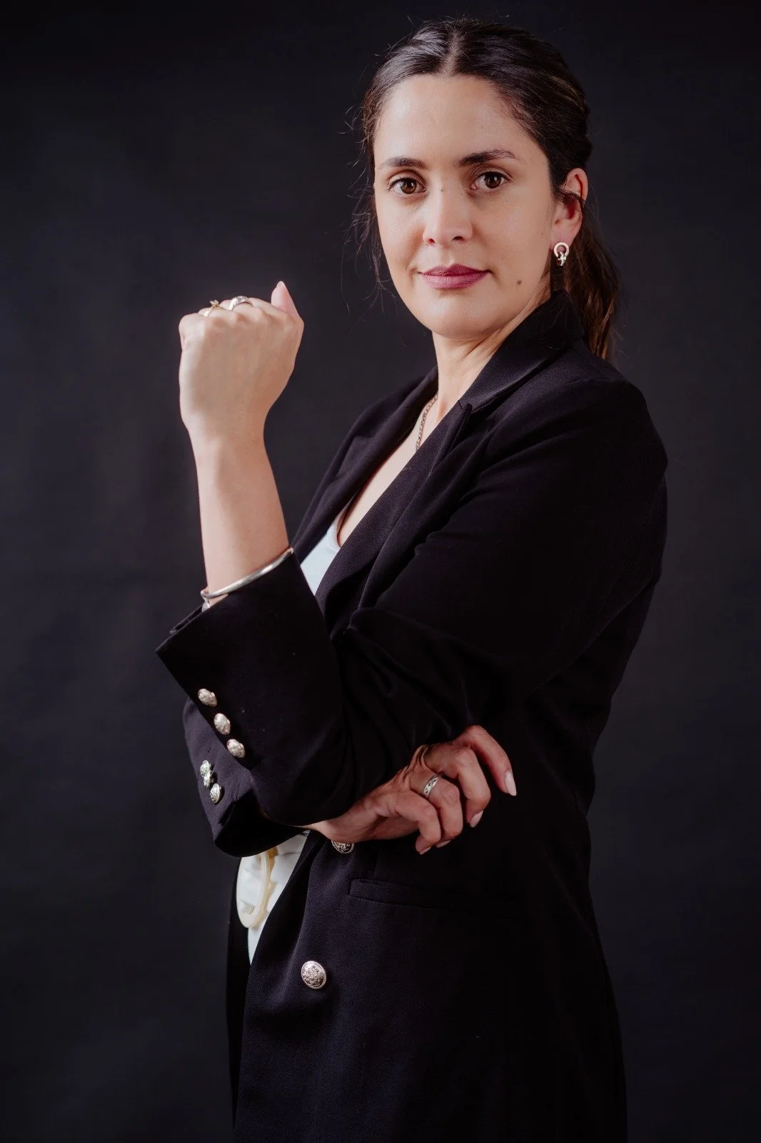 Mujer con expresión decidida, levantando el puño en señal de fuerza y confianza, vestida con saco negro y fondo oscuro.