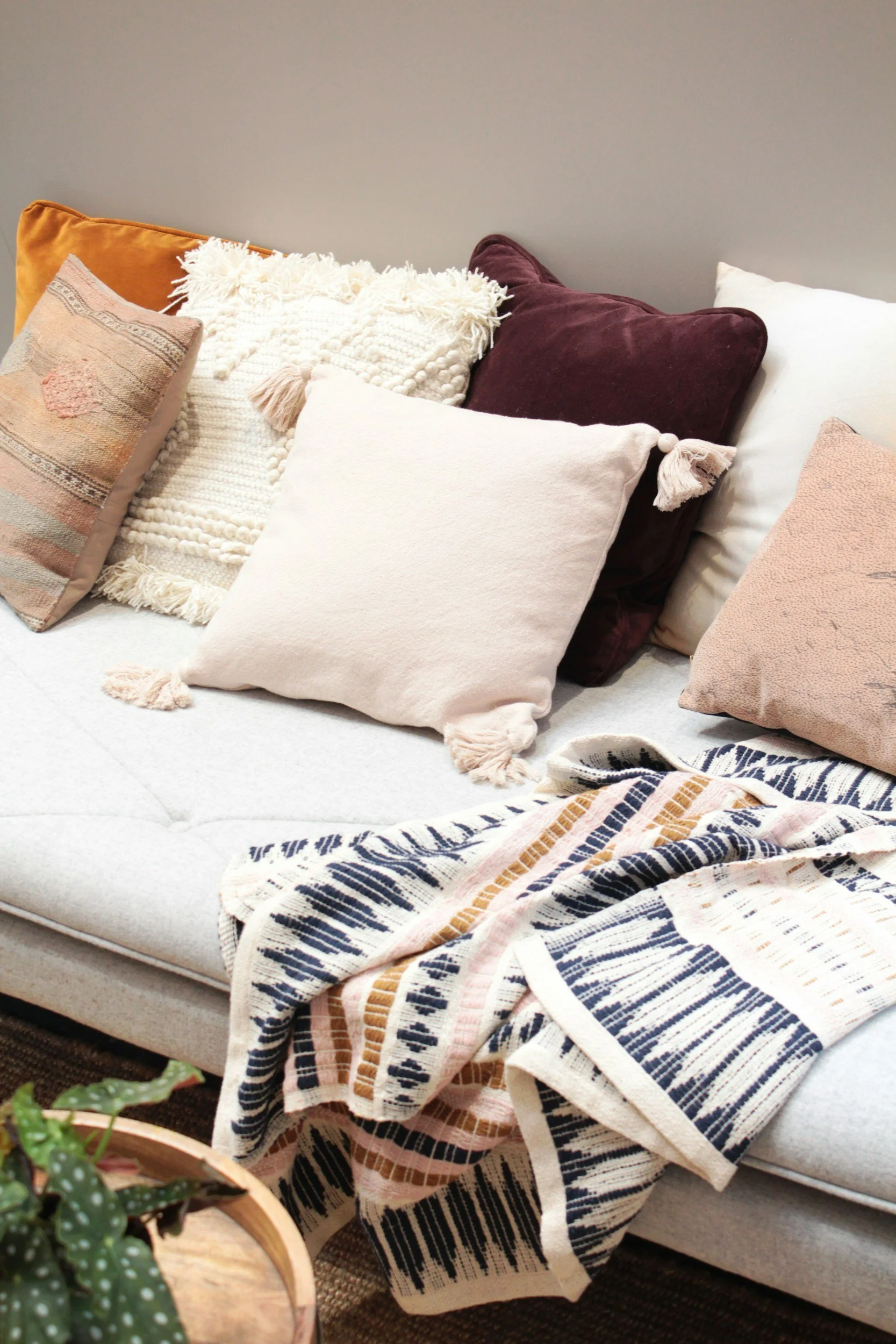 A collection of decorative pillows on a light-colored sofa, including a white pillow with tassels, a maroon velvet pillow, a soft pink pillow, and a patterned pillow with earthy tones, alongside a textured throw blanket with stripes.