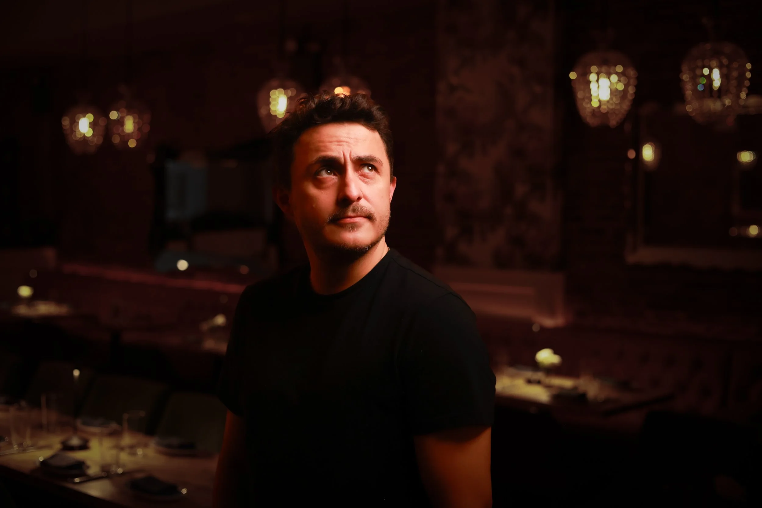 A man with dark hair, wearing a black shirt, looking upward, in a dimly lit restaurant or lounge with ornate lighting fixtures in the background.