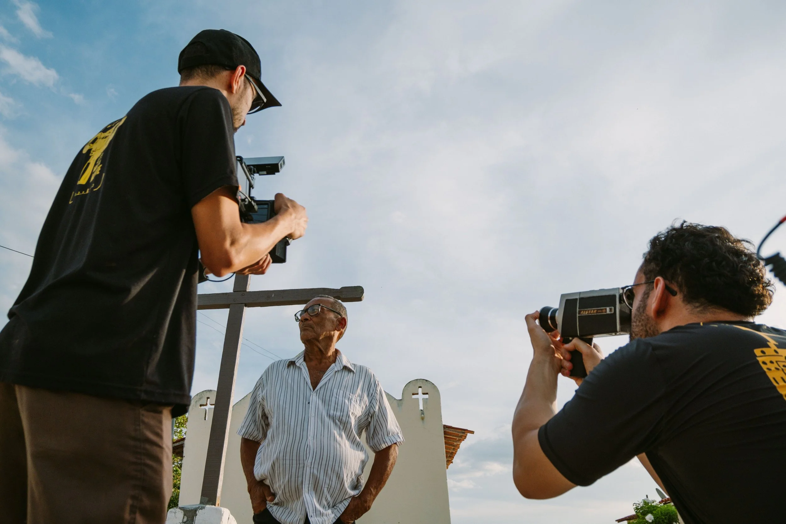 A group of men, two of whom are holding vintage video cameras, film an older man wearing glasses and a striped button-up shirt outdoors against a blue sky with some clouds. The scene appears to be a small filming project or interview.