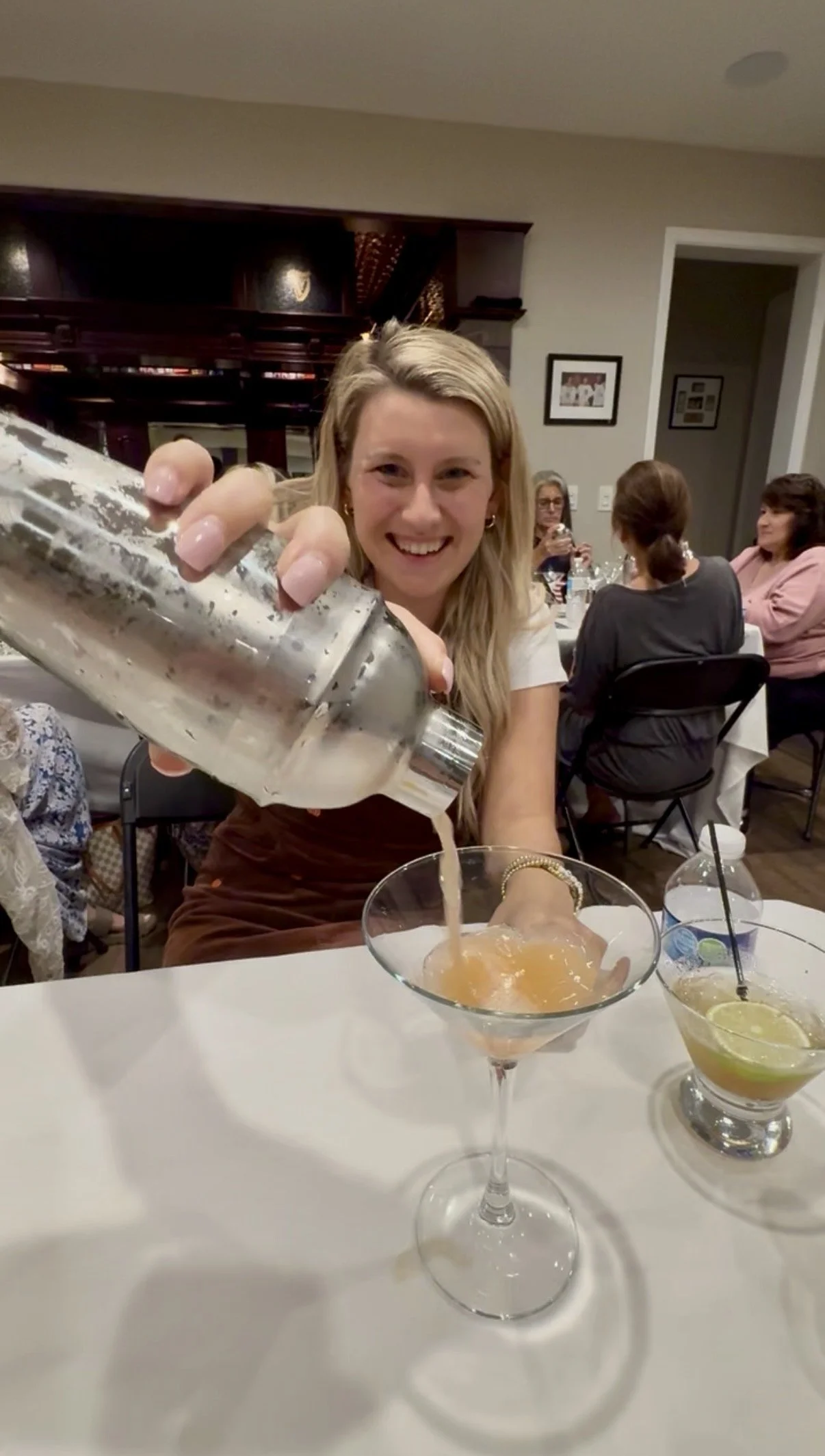 A smiling woman pouring a drink from a shaker into a martini glass at a social gathering, with two other women seated at a table in the background, and two cocktails with lemon slices on the white table in the foreground.