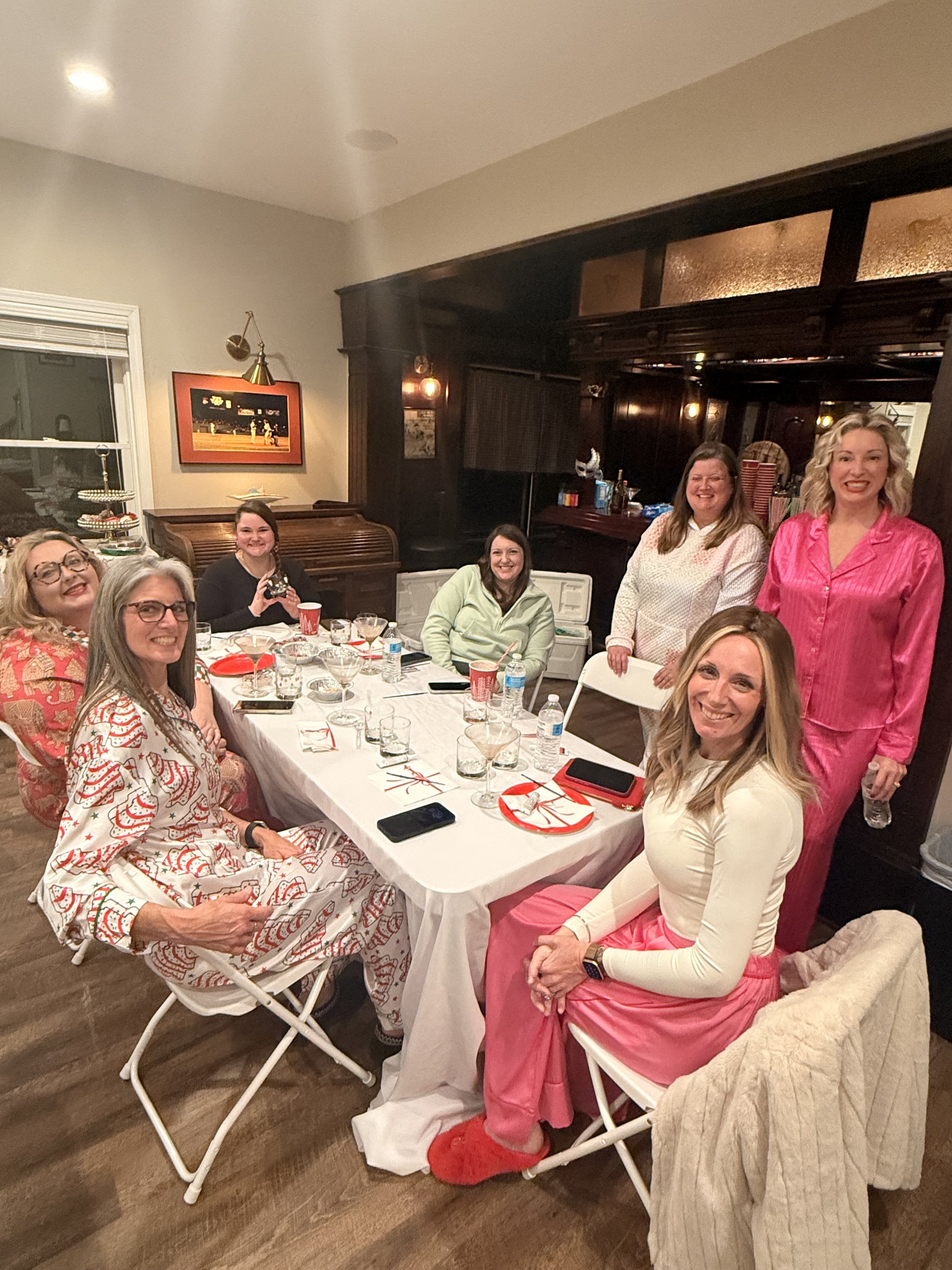 Group of women gathered around a table celebrating a festive occasion, some wearing holiday-themed pajamas and slippers.