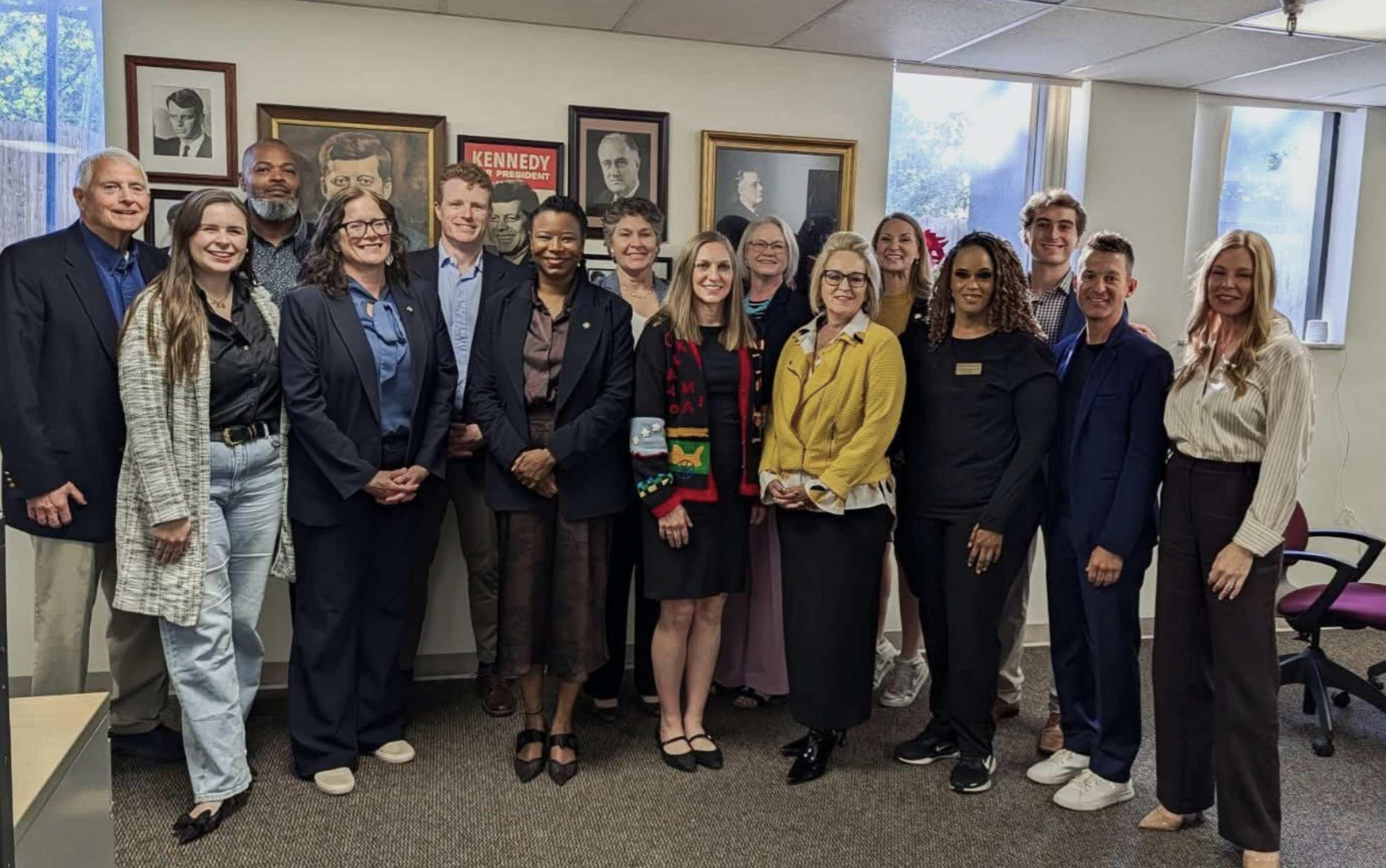 A diverse group of 15 people standing together in an office, smiling for a group photo with framed portraits on the wall behind them.