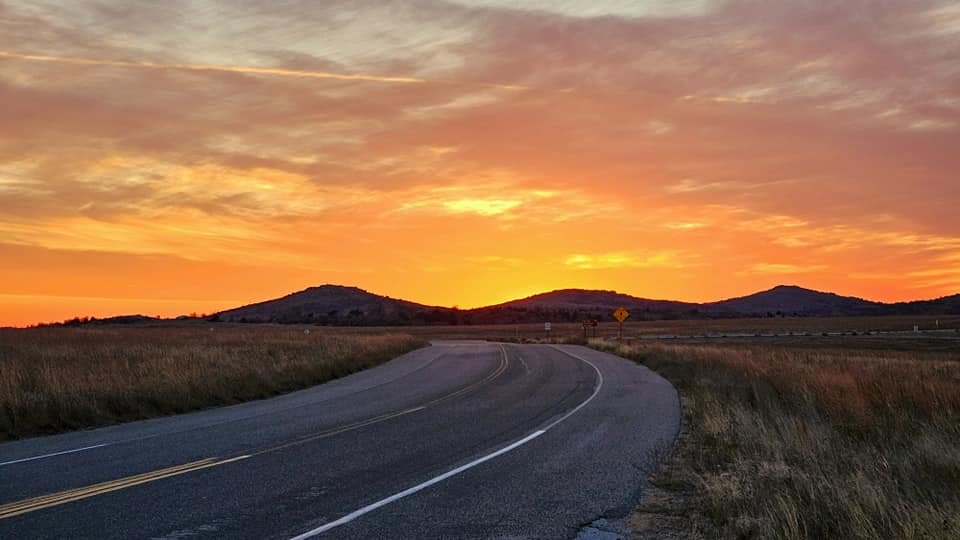 A winding road through grassy fields with hills in the distance during a colorful sunset with orange and yellow hues in the sky.