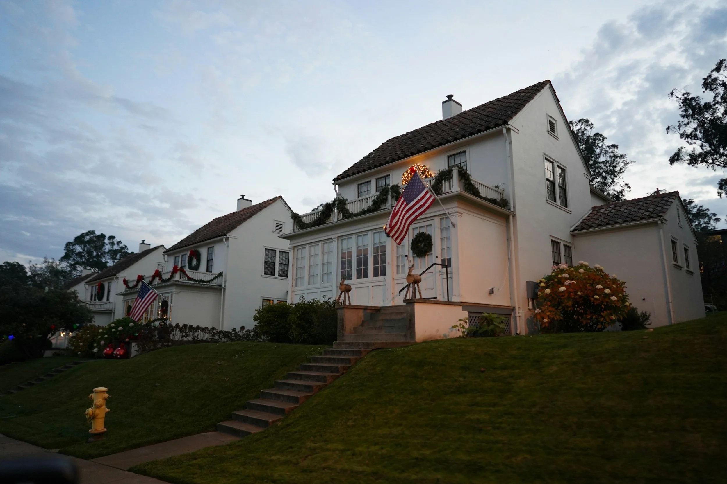 A large white house decorated with Christmas ornaments and American flags. The house has multiple levels with a sloped roof and windows. There are Christmas wreaths, garlands, and lights on the house and yard, with a clear sky and trees in the background.