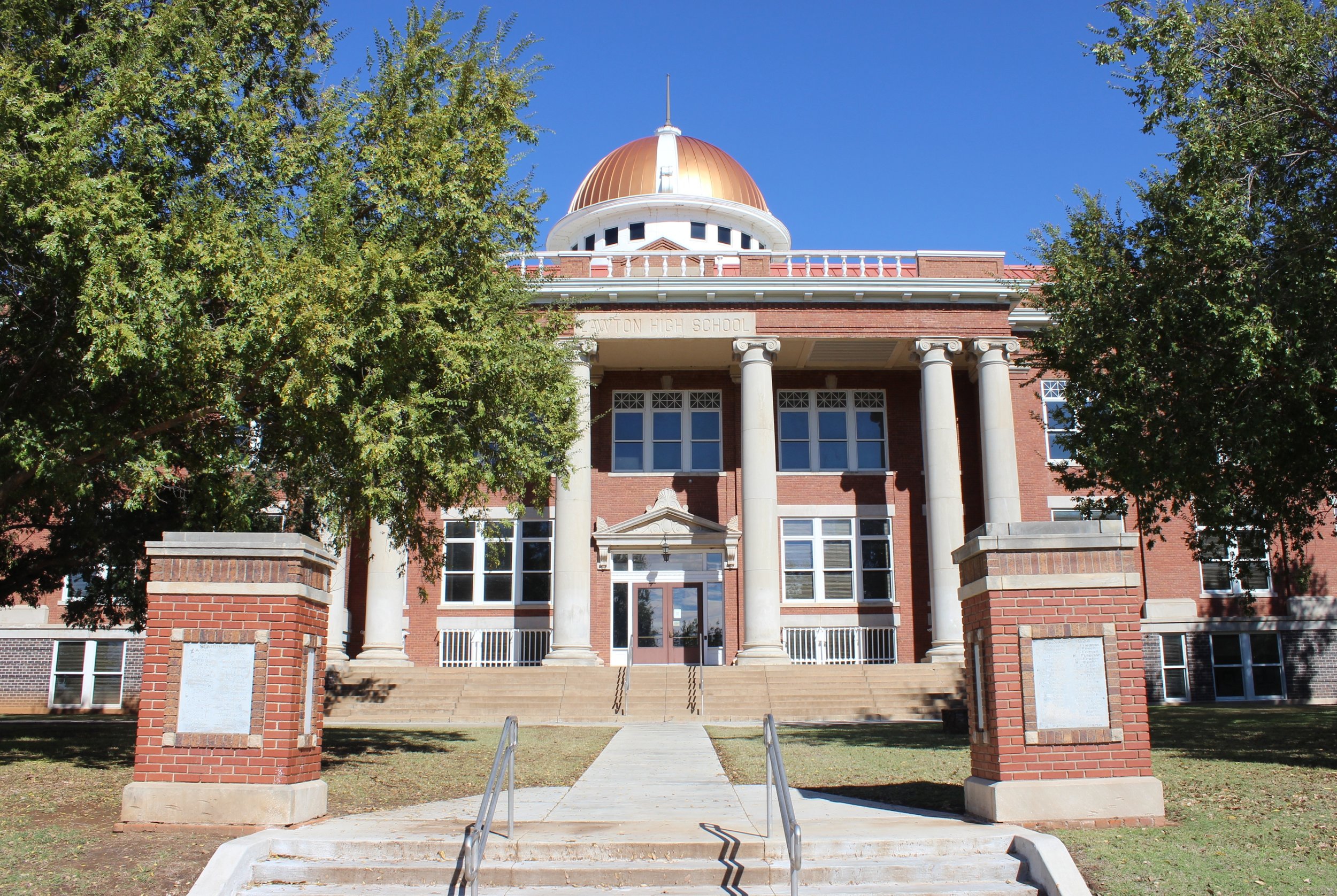 Front view of a Lawton's City Hall, building with large white columns, mannequins, and a rounded copper dome under a clear blue sky, framed by green trees.