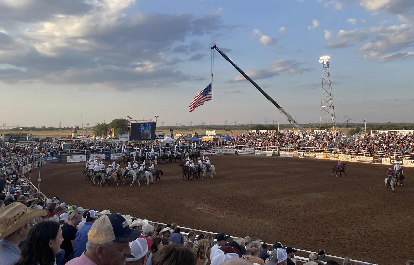 A rodeo event with spectators in the stands, cowboys on horseback on the dirt arena, a large American flag being raised on a crane, and a big screen displaying the event in the background under a partly cloudy sky.