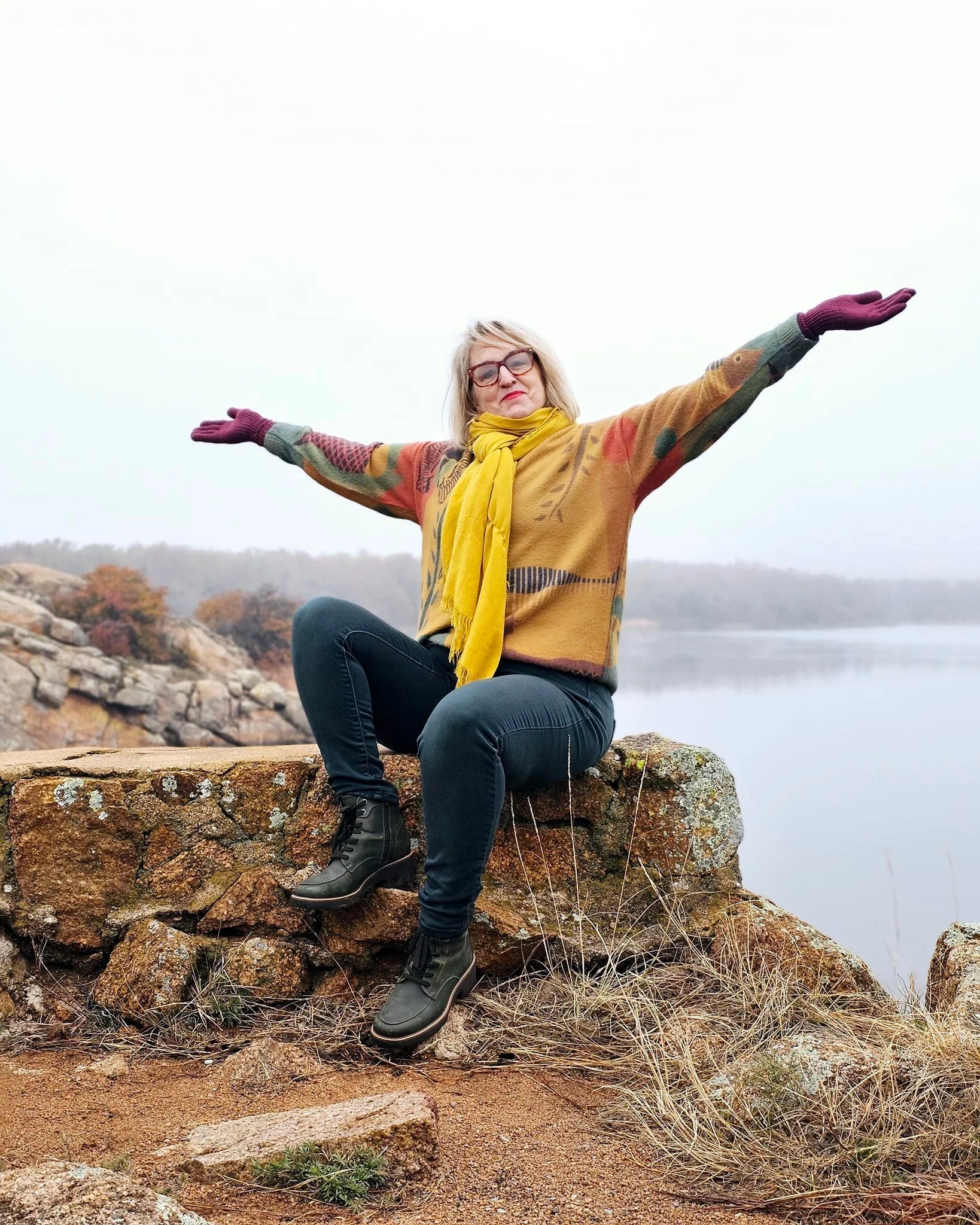 Kathy Suttles is sitting on a rock with her arms outstretched on a cloudy day near a body of water and trees in fall foliage.