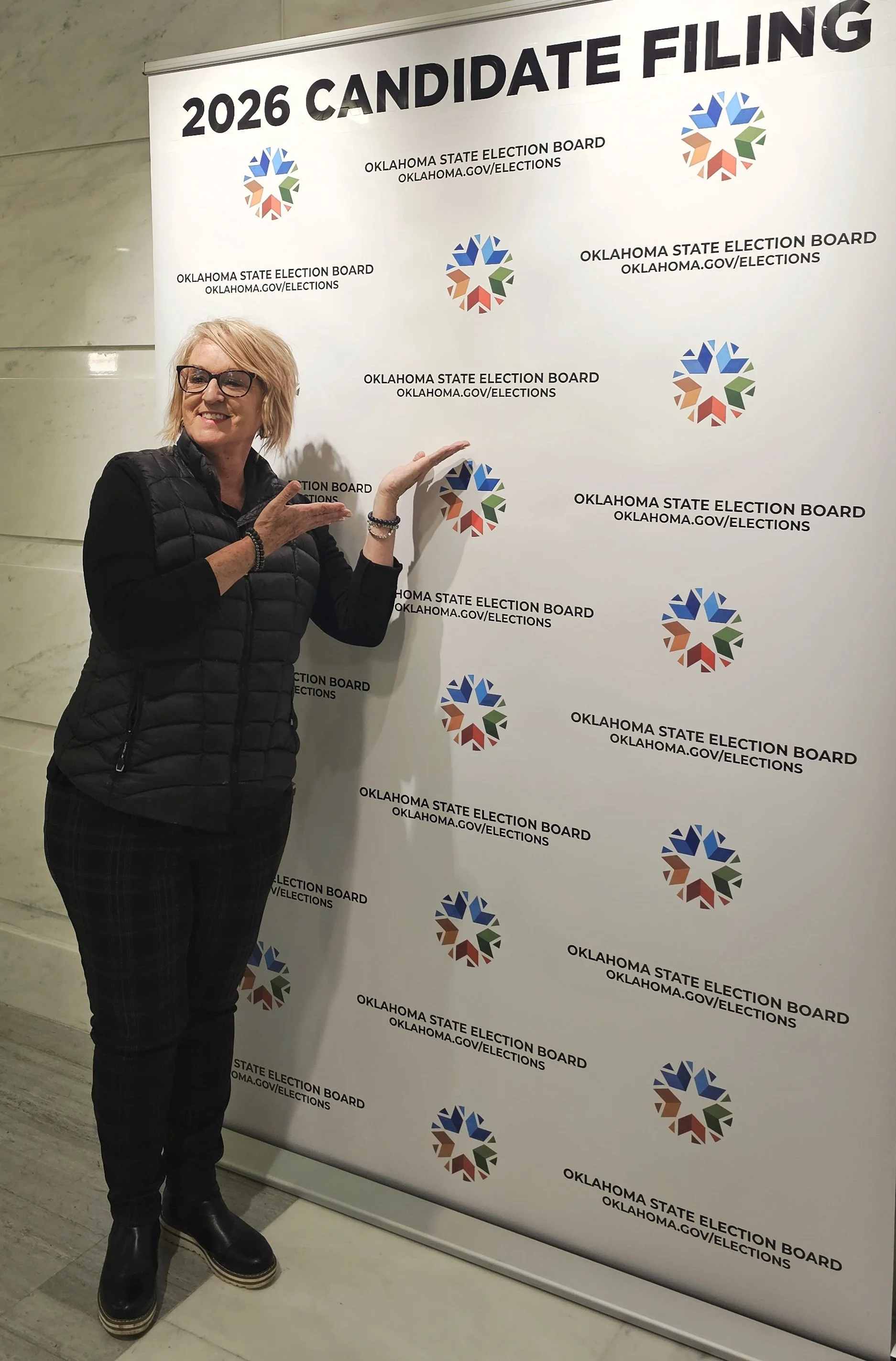 Kathy Suttles standing next to a banner that reads '2026 Candidate Filing' with Oklahoma State Election Board logo and website, smiling and pointing at the logo.