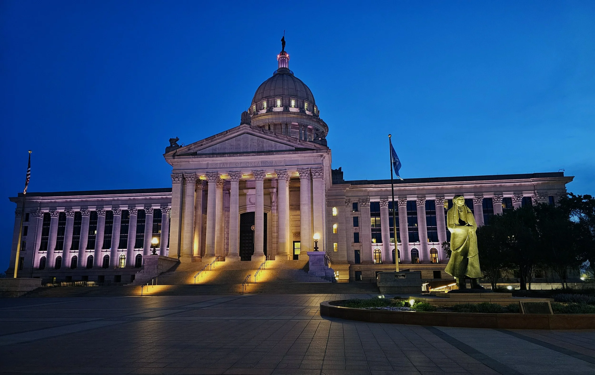 The Oklahoma State Capitol building illuminated at dusk, with a large statue in front and flags flying nearby.