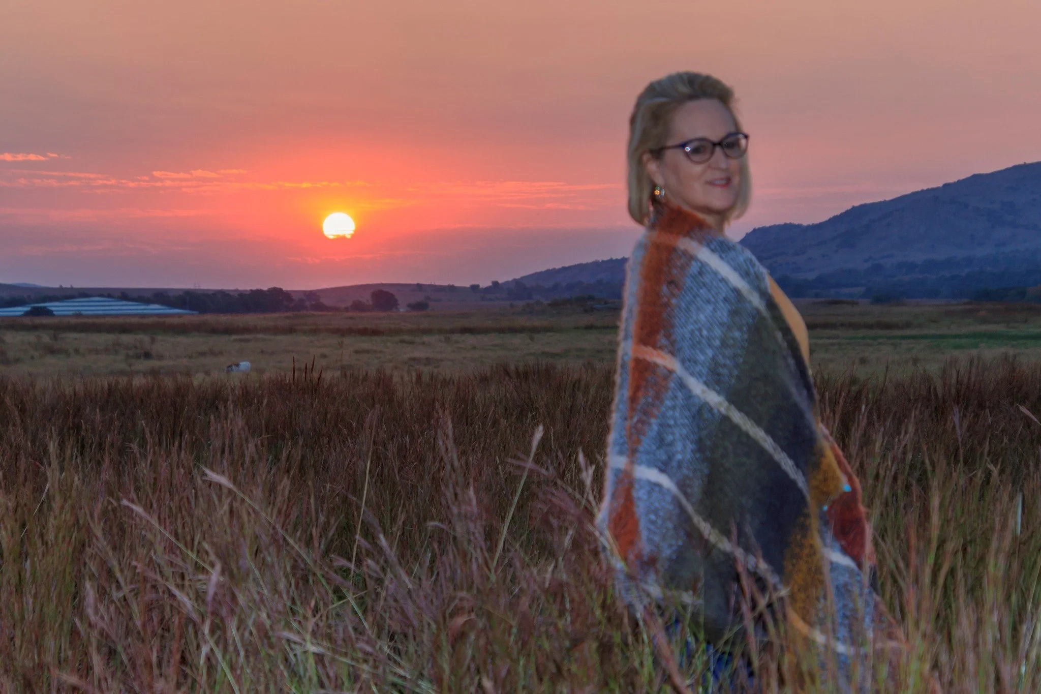 Kathy Suttles smiling while ringing a colorful blanket, standing outdoors in a field at sunset with rolling hills and a pastel sky in the background.