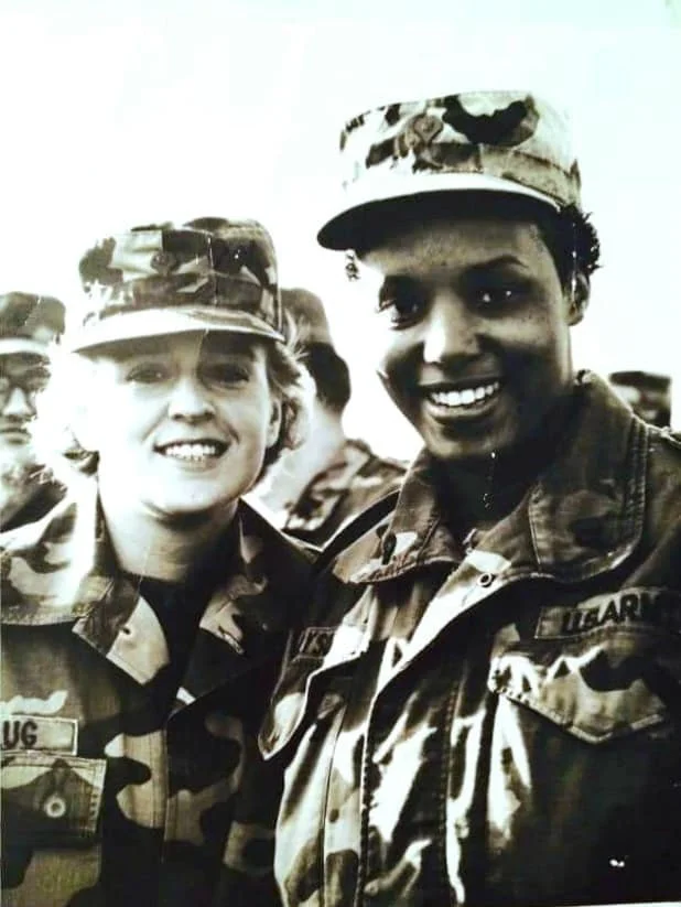 Two soldiers in camouflage uniforms smiling, one is a woman with curly hair, wearing a patrol cap, and the other is a man with short hair, also wearing a military cap, with several other soldiers blurred in the background.