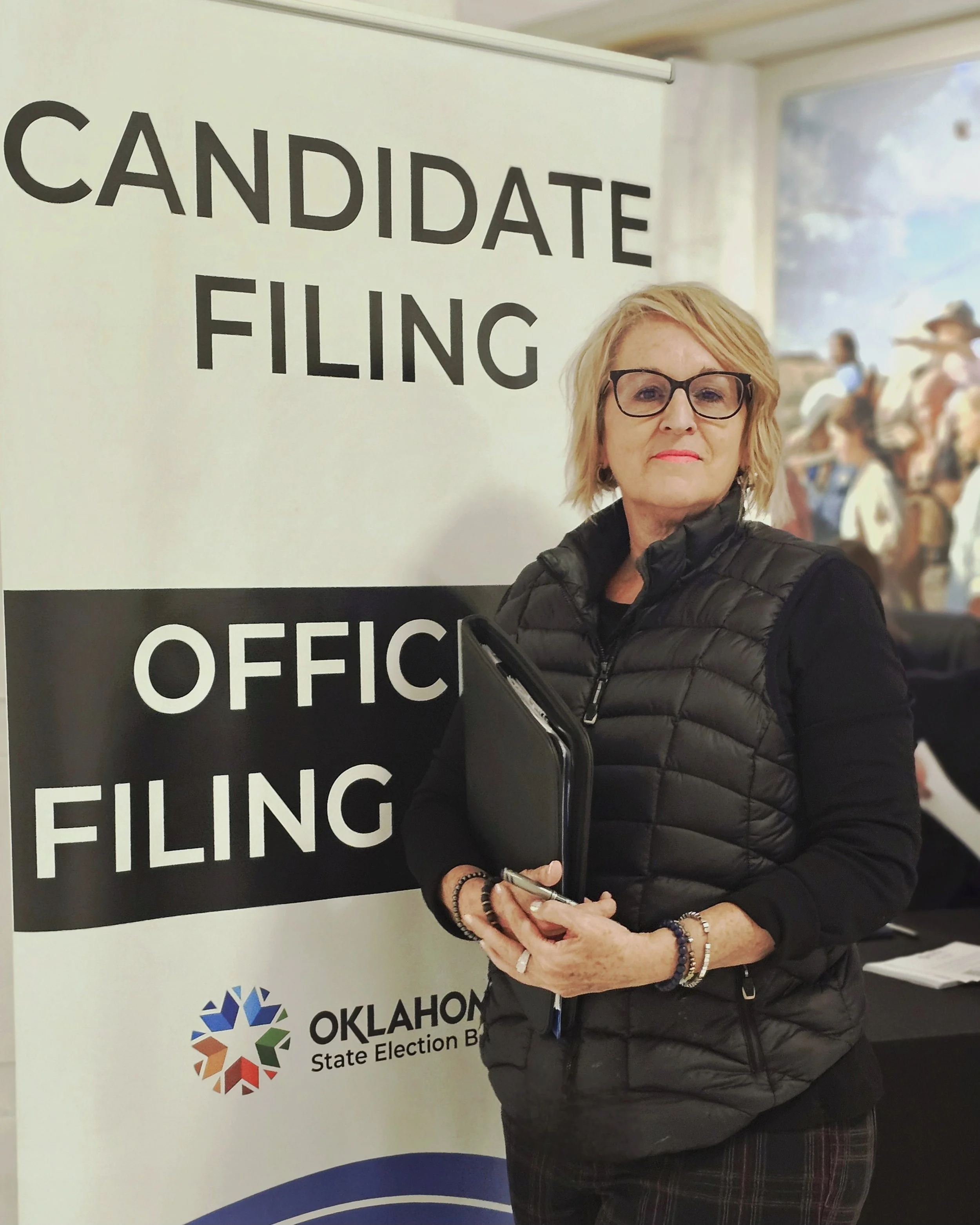 A woman with blonde hair, glasses, and a black puffer jacket standing in front of a sign for an election office, holding a notebook and a pen.