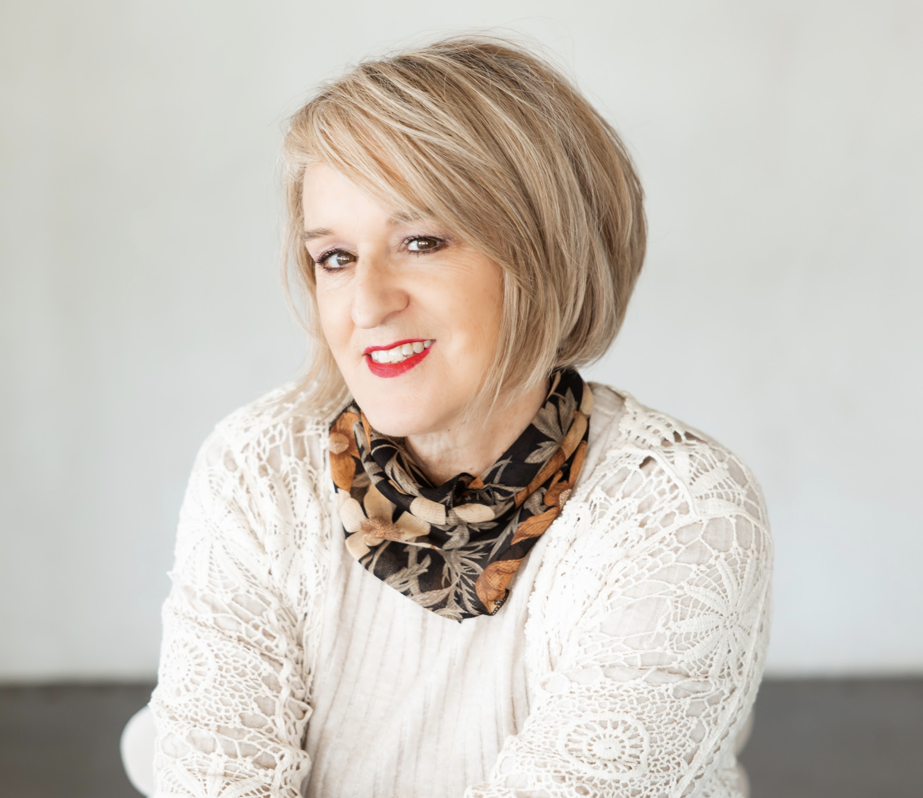 Kathy Suttles - a woman with short, blonde hair, red lipstick, wearing a white lace top and a patterned scarf, smiling softly with arms crossed.
