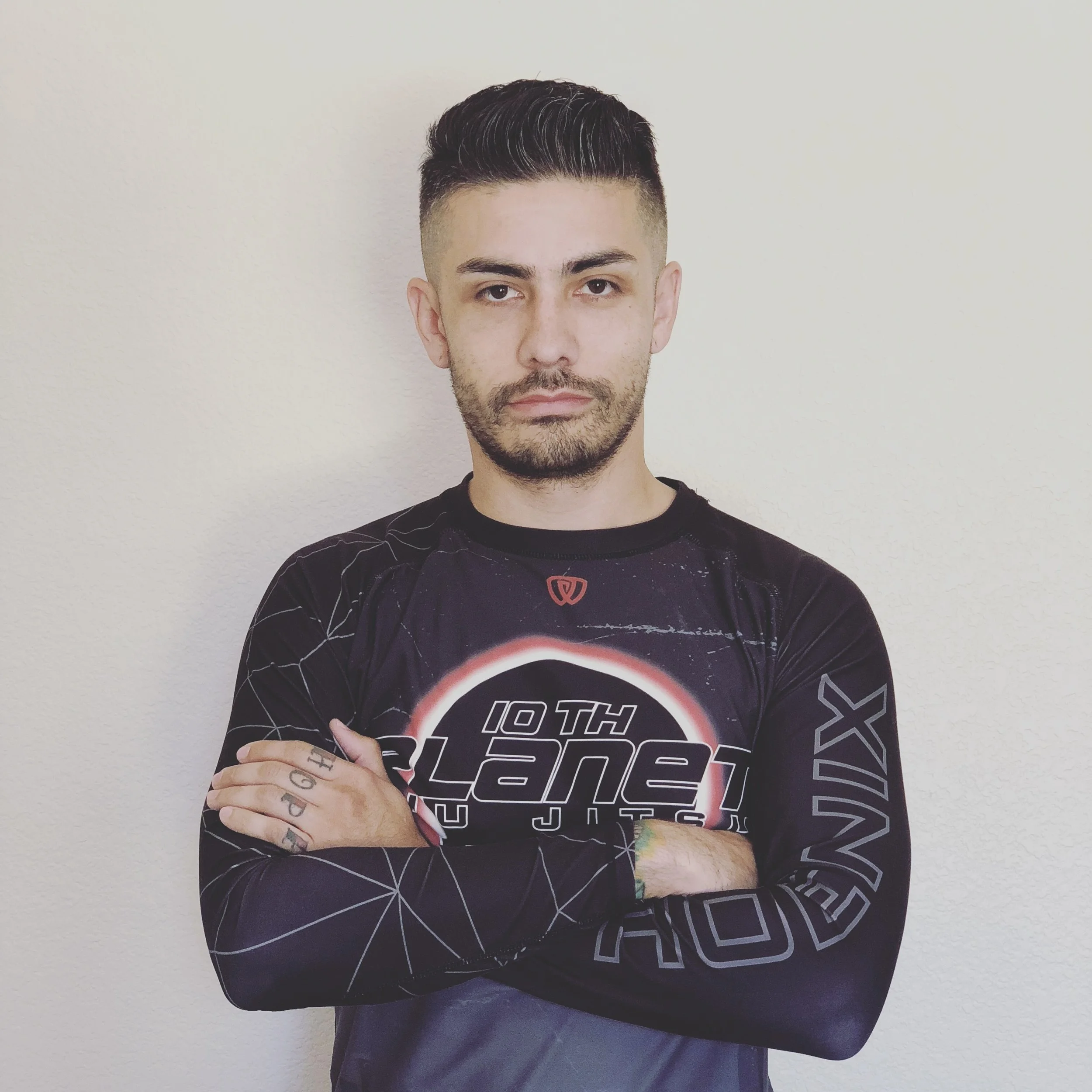 A man with dark hair and tattoos on his fingers, wearing a black shirt with a graphic design, is standing against a plain white wall. His arms are crossed, and he is looking directly at the camera.