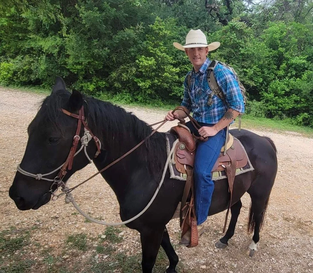 A man in a cowboy hat, plaid shirt, and jeans sitting on a black horse with a brown saddle, on a dirt trail with green trees in the background.