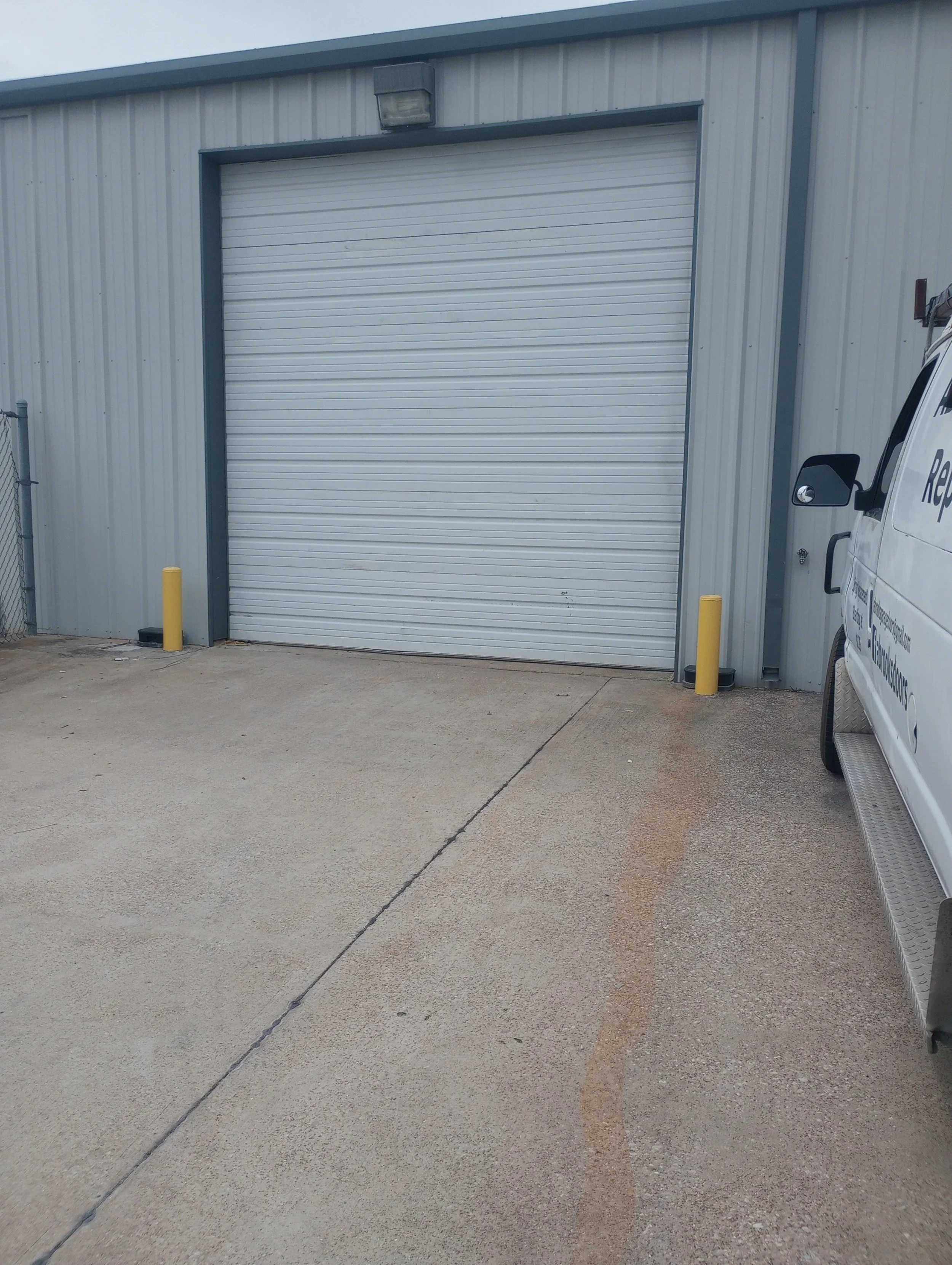 Exterior view of a metal warehouse with a large closed roll-up door, two yellow protective bollards in front, and a white van parked nearby. The ground is concrete.