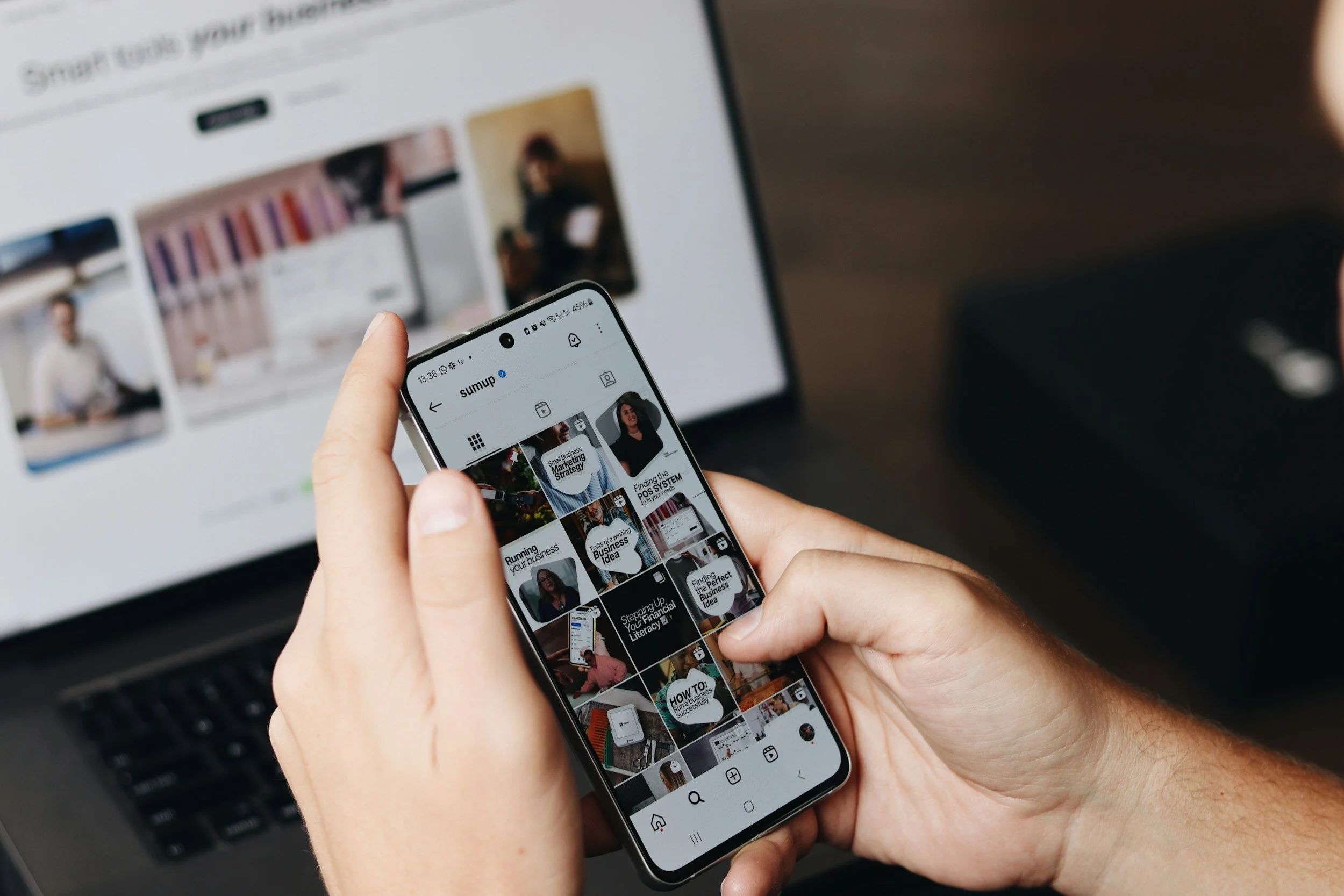 Person holding a smartphone showing a social media profile, with a computer screen in the background displaying a website or online content.