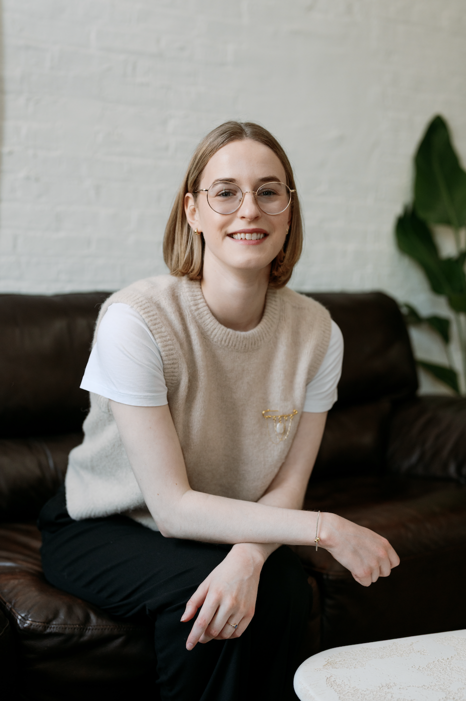 A young woman with short blonde hair, glasses, and a beige sweater vest over a white t-shirt sitting on a dark brown leather couch in a room with a white brick wall and a large green plant.
