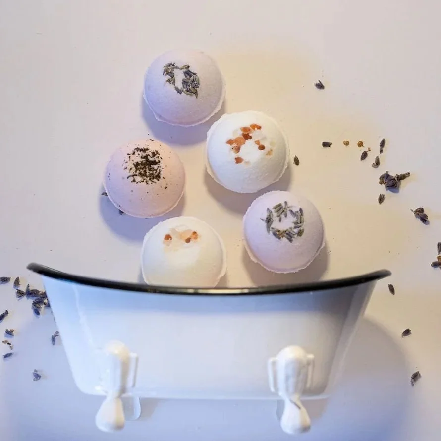 Five bath bombs with different colors and textures, surrounded by scattered lavender flowers on a white surface, viewed from above.